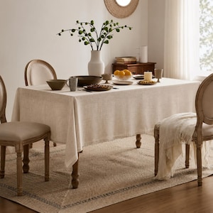 Puede incluir: Una mesa de comedor rectangular cubierta con un mantel de lino beige texturizado, rodeada por cuatro sillas de madera con asientos tapizados. La mesa está dispuesta con cuencos de cerámica, platos, un jarrón blanco y un cuenco con naranjas.