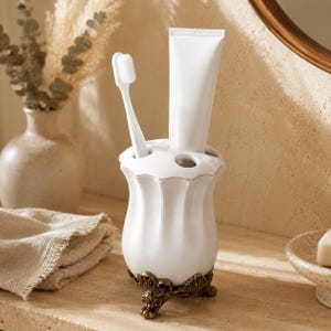 Peut inclure: Un porte-brosse à dents en céramique blanche avec un corps cannelé et festonné, reposant sur une base en métal ornée de couleur bronze antique à trois pieds. Il contient une brosse à dents blanche et un tube de dentifrice blanc vertical.