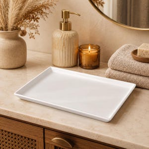 Peut inclure: Un plateau rectangulaire en céramique blanche avec une finition lisse et brillante et des bords légèrement surélevés. Le plateau est posé sur une surface plane devant un distributeur de savon beige, une bougie allumée et deux serviettes.