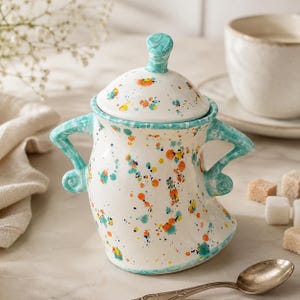May include: A white ceramic sugar bowl with a lid, featuring two curved handles and a colourful paint splatter pattern in shades of turquoise, orange, and yellow. The bowl has a rounded body and a matching lid with a small knob handle on top.