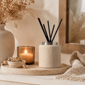May include: A cylindrical beige stone diffuser vessel with a silver-toned metal collar and five black reed sticks inserted. The diffuser sits on a circular travertine tray alongside a small amber glass candle holder with a lit candle.