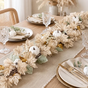 Peut inclure: Un chemin de table composé de feuilles d'érable couleur crème, de petites citrouilles blanches et de baies blanches sur un chemin de table en lin beige. La table est dressée avec des assiettes blanches, des couverts dorés et des verres en verre.