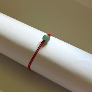 May include: A delicate bracelet featuring a single smooth, round, light green gemstone bead centered on a thin, vibrant red cord. The cord is secured with a small, intricate knot on either side of the bead, creating a simple and minimalist design.