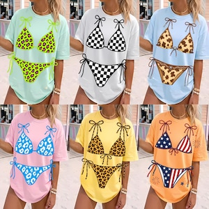 May include: A collection of six t-shirts, each featuring a different bikini design. The designs include leopard print, checkered patterns, and patriotic themes. The shirts are in various colors, including light blue, white, pink, yellow, and orange.