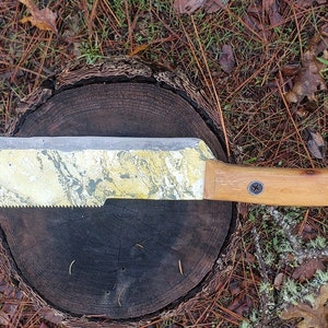May include: A large machete with a curved blade and a wooden handle. The blade has a serrated edge and a gold and silver finish. The handle is attached with two black screws. The machete is resting on a tree stump.
