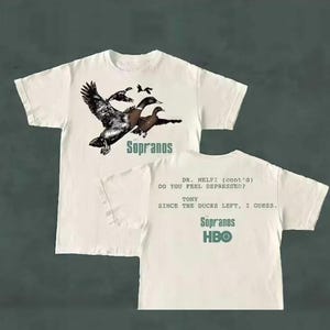 May include: White t-shirt featuring a graphic of ducks in flight with the text "Sopranos." The back of the shirt displays quotes from the show and the HBO logo. A classic tee for fans of the series.
