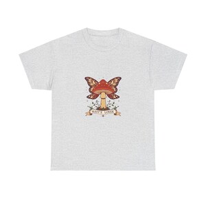 Könnte beinhalten: Hellgraues T-Shirt mit einem fantasievollen Design. Das Design zeigt einen Pilz mit Schmetterlingsflügeln in Rot-, Braun- und Goldtönen. Der Text "MASTE LOEEK" ist auf einem Banner darunter gedruckt.