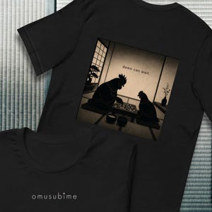 Op de afbeelding: Een zwart t-shirt met een grafische print van twee kippen die een bordspel spelen in een kamer met tatami-matten en een bonsaiboom. De tekst "dawn can wait." staat erboven en "omusubime" staat op de onderste voorzijde.