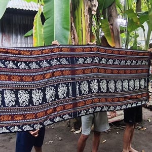 Pode incluir: Um tecido retangular com um padrão detalhado. O tecido apresenta um fundo azul escuro com motivos florais e de elefantes brancos repetidos, delimitados por faixas laranja e marrom. O tecido é segurado por várias pessoas.
