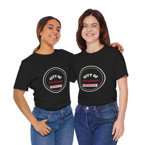 Op de afbeelding: Een zwart t-shirt met korte mouwen en een cirkelvormige witte afbeelding met de tekst CITY OF GOOD NEIGHBORS en BUFFALO NY in rood en wit. Het ontwerp bevat een witte cirkelvormige rand en een rood banier-element in het midden.