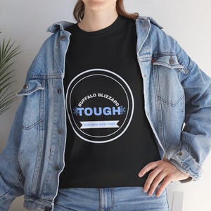 Op de afbeelding: Een zwart t-shirt met een witte cirkelvormige afbeelding met de tekst BUFFALO BLIZZARD TOUGH en WESTERN NEW YORK, geaccentueerd door lichtblauwe sneeuwvlokpictogrammen, gedragen onder een lichtblauw spijkerjack met metalen knopen en spijkerbroek.