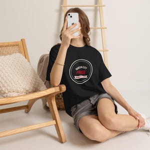 Op de afbeelding: Een zwart t-shirt met korte mouwen en een cirkelvormige witte afbeelding op de borst. De tekst in de cirkel luidt QUEEN CITY PRIDE in wit en rood, met daaronder een banner met de tekst BUFFALO NY CLASSIC in witte letters op een rode achtergrond.