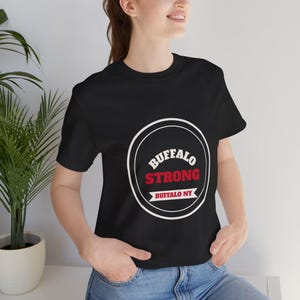 Op de afbeelding: Een zwart t-shirt met korte mouwen voorzien van een witte cirkelvormige afbeelding met de tekst BUFFALO in het wit, STRONG in het rood, en een banner met de tekst BUFFALO NY in het rood. Het ontwerp is gecentreerd op de borst.