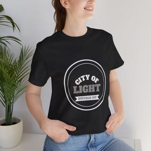 Op de afbeelding: Een zwart t-shirt met korte mouwen en een witte cirkelvormige afbeelding op de borst. De afbeelding bevat de tekst CITY OF LIGHT in wit en grijs, met de woorden BUFFALO NY in het wit op een banner onderaan de cirkel.