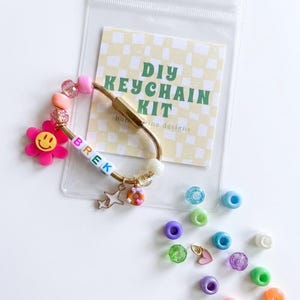 May include: A DIY keychain kit in a clear plastic bag with a card labelled DIY KEYCHAIN KIT. The kit includes a gold-tone oval carabiner, letter beads spelling BREK, a pink flower charm with a smiley face, a star charm, and a donut charm. Loose beads in blue, green, purple, orange, and white, plus a small pink heart charm are included.