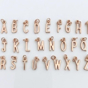 May include: A collection of twenty-six individual rose gold-toned metal letter charms, arranged in three rows from A to Z. Each letter features a small circular jump ring at the top for attachment, showcasing a polished finish and sans-serif font.