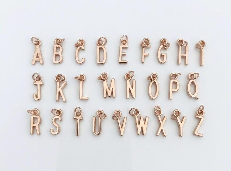 May include: A collection of twenty-six individual rose gold-toned metal letter charms, arranged in three rows from A to Z. Each letter features a small circular jump ring at the top for attachment, showcasing a polished finish and sans-serif font.