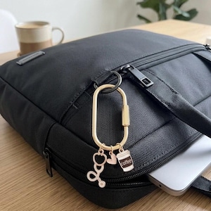 May include: A gold-toned oval carabiner keyring attached to a black fabric bag. The keyring features three small charms: a rose gold stethoscope, a small rose gold heart, and a white and brown coffee cup charm with the word coffee printed on it.