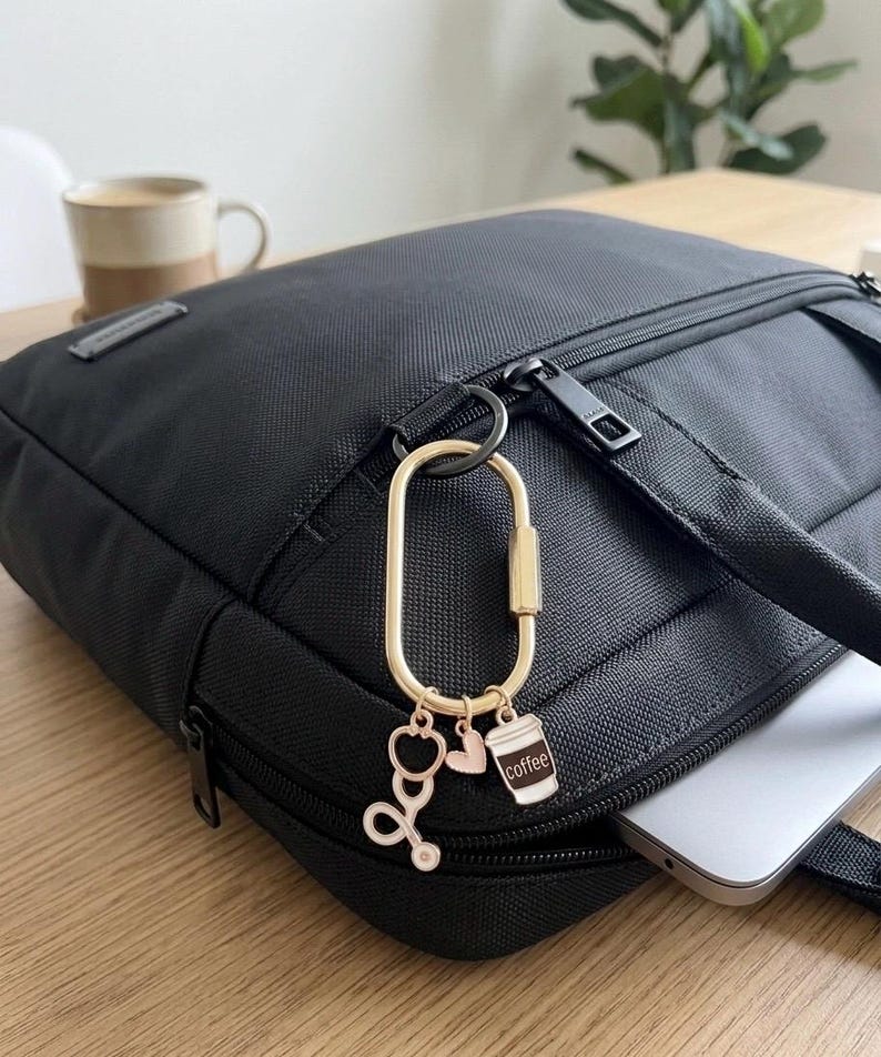 May include: A gold-toned oval carabiner keyring attached to a black fabric bag. The keyring features three small charms: a rose gold stethoscope, a small rose gold heart, and a white and brown coffee cup charm with the word coffee printed on it.