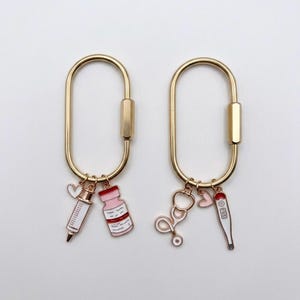May include: Two gold-toned oval carabiner keychains. The left keychain features a white and pink syringe charm, a pink medicine vial charm, and a small white heart. The right keychain features a gold stethoscope charm, a pink heart, and a thermometer.