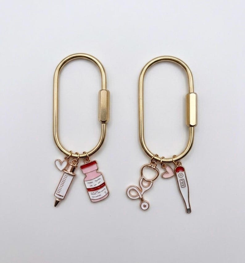 May include: Two gold-toned oval carabiner keychains. The left keychain features a white and pink syringe charm, a pink medicine vial charm, and a small white heart. The right keychain features a gold stethoscope charm, a pink heart, and a thermometer.