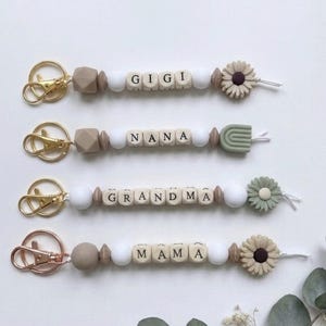 May include: Four keychains featuring gold or rose gold lobster clasps, silicone beads in beige, white, and brown, and lettered cubes spelling GIGI, NANA, GRANDMA, and MAMA. Each keychain ends with a silicone flower or rainbow charm.