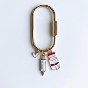 May include: A gold-toned oval carabiner keyring featuring three dangling enamel charms: a small white heart with a gold border, a white syringe with gold accents, and a light pink medicine vial with a dark red cap and gold-toned trim.
