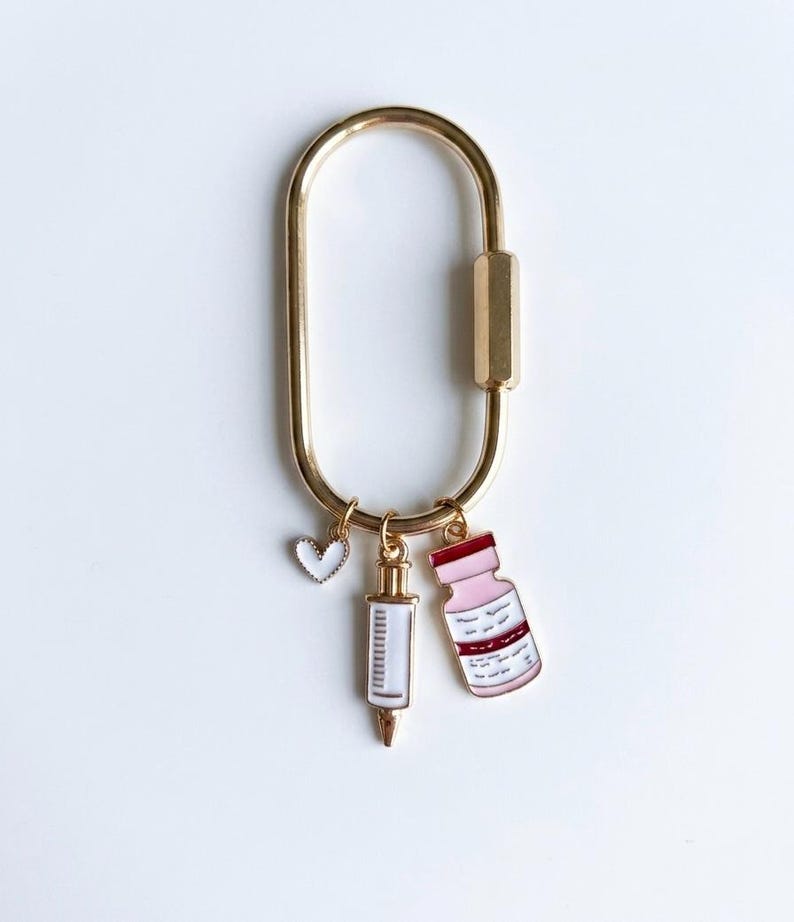 May include: A gold-toned oval carabiner keyring featuring three dangling enamel charms: a small white heart with a gold border, a white syringe with gold accents, and a light pink medicine vial with a dark red cap and gold-toned trim.