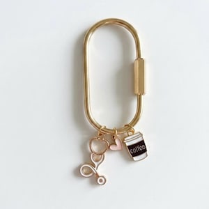 May include: A gold-toned oval carabiner keyring featuring three dangling enamel charms: a white stethoscope, a small light pink heart, and a white coffee cup with a dark brown band and the word coffee printed in white lettering on the front.