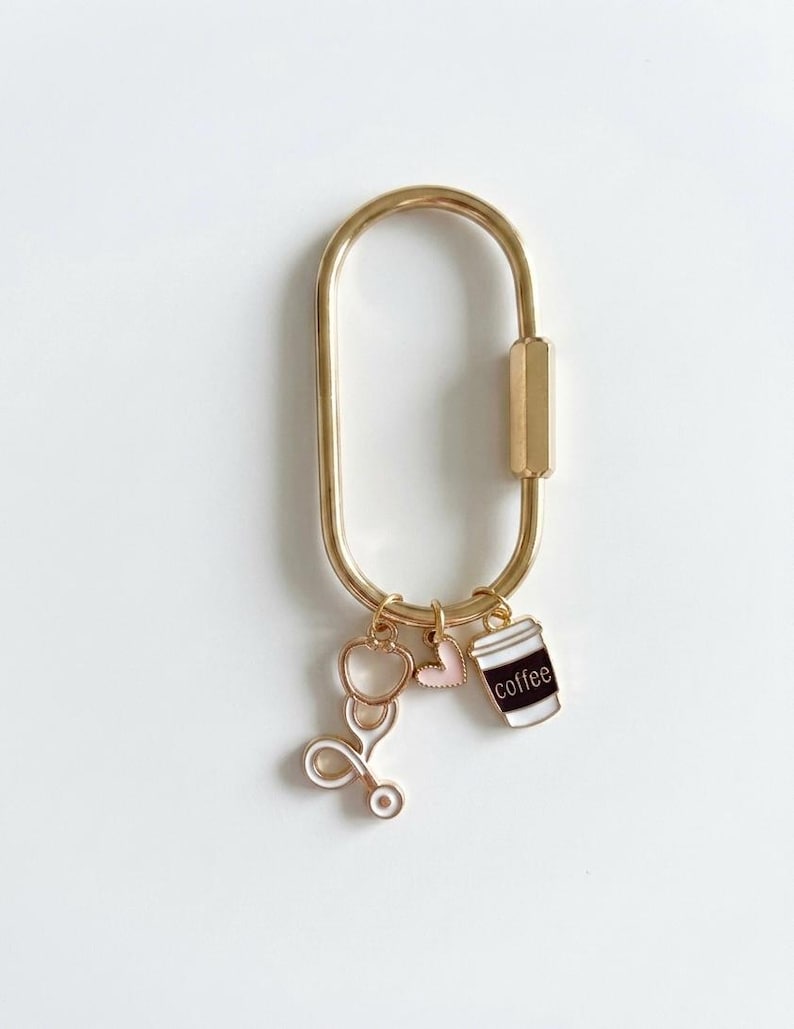 May include: A gold-toned oval carabiner keyring featuring three dangling enamel charms: a white stethoscope, a small light pink heart, and a white coffee cup with a dark brown band and the word coffee printed in white lettering on the front.