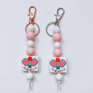 May include: Two keychains featuring round silicone beads in white and light pink. Each keychain includes a square white bead with a graphic of two pink pickleball paddles and an orange ball, with the text PICKLEBALL printed in green and pink.