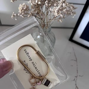 May include: A gold-toned metal carabiner keyring attached to a white display card labelled "honeytwine designs". The keyring features three small charms: a white and gold stethoscope, a small pink heart, and a black and white coffee cup charm.