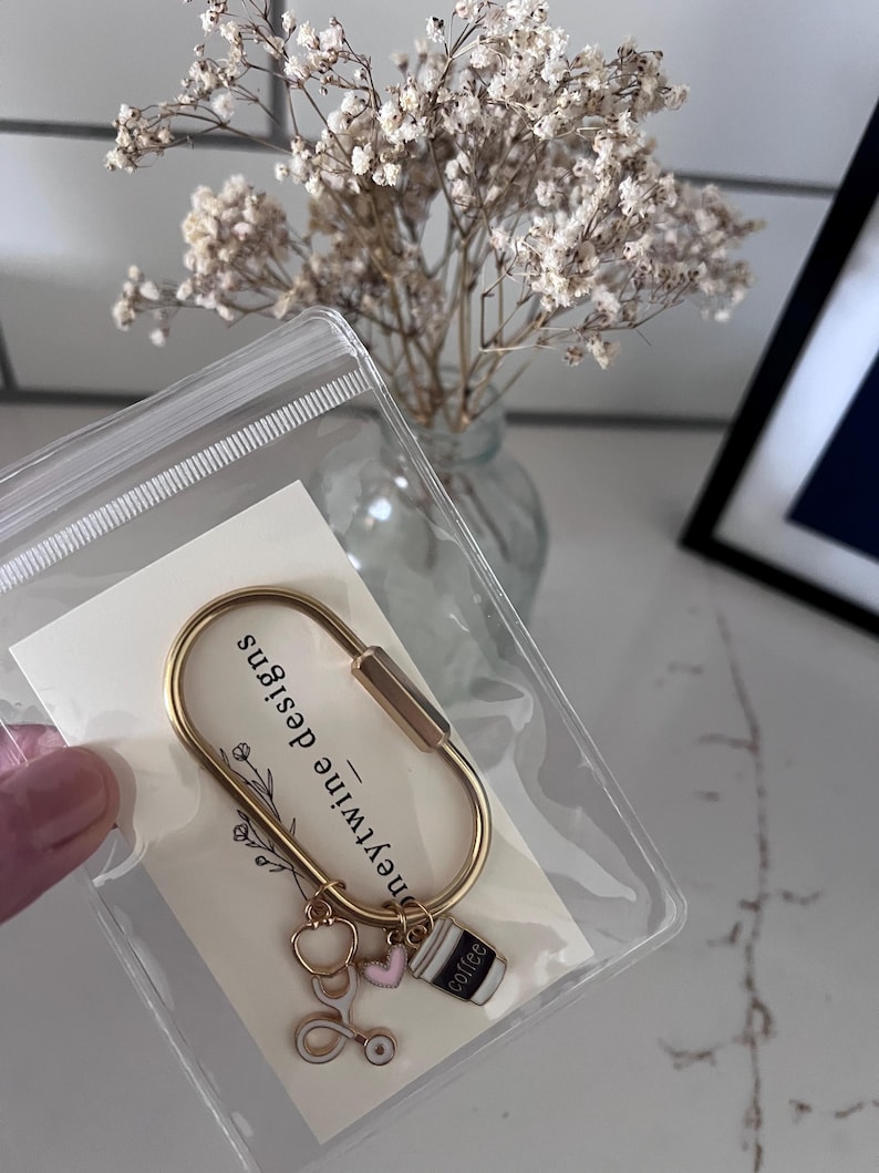 May include: A gold-toned metal carabiner keyring attached to a white display card labelled "honeytwine designs". The keyring features three small charms: a white and gold stethoscope, a small pink heart, and a black and white coffee cup charm.