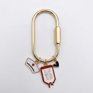 May include: A gold-toned oval carabiner keyring featuring three dangling enamel charms: a white nurse cap with a red cross, a red and white blood bag with black lines, and a small black heart with a gold-toned beaded edge border.