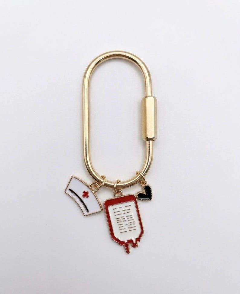 May include: A gold-toned oval carabiner keyring featuring three dangling enamel charms: a white nurse cap with a red cross, a red and white blood bag with black lines, and a small black heart with a gold-toned beaded edge border.
