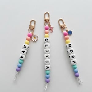 May include: Three personalised keychains featuring colourful silicone beads in pastel shades of pink, yellow, blue, and purple. Each keychain has white square letter beads spelling the names AVA, OLIVIA, and NORA, with gold-tone metal clasps and charms.