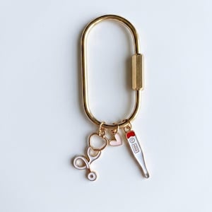 May include: A gold-toned oval carabiner keyring featuring three dangling charms: a white and gold stethoscope, a small light pink heart, and a white thermometer charm with a red top and black markings, all attached by small gold jump rings.