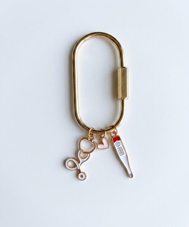 May include: A gold-toned oval carabiner keyring featuring three dangling charms: a white and gold stethoscope, a small light pink heart, and a white thermometer charm with a red top and black markings, all attached by small gold jump rings.