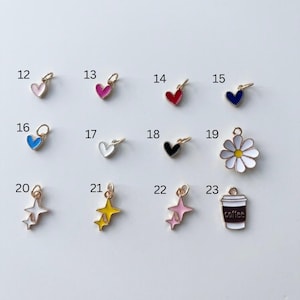 May include: A collection of twelve small metal charms labelled 12 through 23. The charms include various heart shapes in pink, red, blue, white, and black, a white daisy with a yellow centre, star clusters in white, yellow, and pink, and a coffee cup.