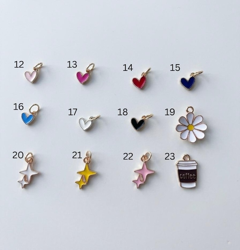 May include: A collection of twelve small metal charms labelled 12 through 23. The charms include various heart shapes in pink, red, blue, white, and black, a white daisy with a yellow centre, star clusters in white, yellow, and pink, and a coffee cup.