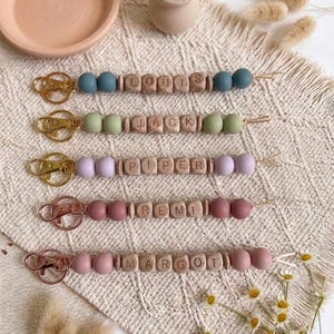 May include: Five personalized keychains featuring gold-toned metal clasps, smooth wooden letter beads spelling the names LOUIS, JACK, PIPER, REMI, and MARGOT, and round silicone beads in shades of blue, sage green, lavender, mauve, and dusty rose.