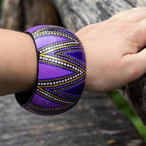 Puede incluir: Un brazalete ancho y morado con un patrón geométrico de puntos y líneas blancos, dorados y negros. El brazalete se lleva en un brazo, apoyado sobre una superficie de madera. El diseño del brazalete es simétrico y llamativo.