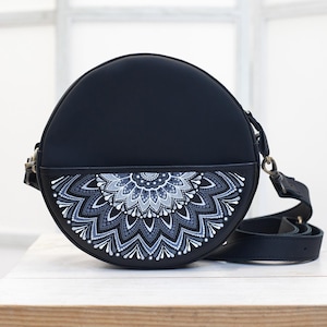 May include: A black, round crossbody bag with a decorative mandala design in shades of gray and white. The bag has a black adjustable strap and a small pocket on the front. The bag is sitting on a white surface.