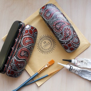 Two eyeglass cases, hand-painted with red and silver paisley design. View from the top on the envelope with the brand logo, showing materials used to paint cases.