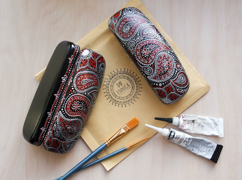 Two eyeglass cases, hand-painted with red and silver paisley design. View from the top on the envelope with the brand logo, showing materials used to paint cases.
