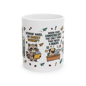 May include: A white ceramic mug featuring colorful cartoon illustrations of a raccoon holding a folder and a pug sitting at a desk. The text reads: Workin' hard, or hardly workin'? and When your computer isn't the only thing that needs a reboot.