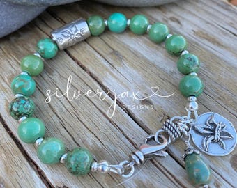May include: A turquoise beaded bracelet with a silver clasp and charm. The bracelet features round, green-blue beads and silver accents. A silver charm with a floral design hangs from the clasp. The text "silverjax designs" is visible.