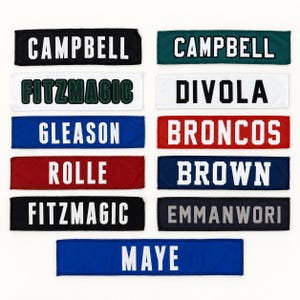 May include: A collection of eleven rectangular fabric name patches in various colors including black, teal, white, blue, red, and dark blue. Each patch features bold, block-style lettering displaying names and words such as CAMPBELL, FITZMAGIC, DIVOLA, GLEASON, BRONCOS, ROLLE, BROWN, EMMANWORI, and MAYE.