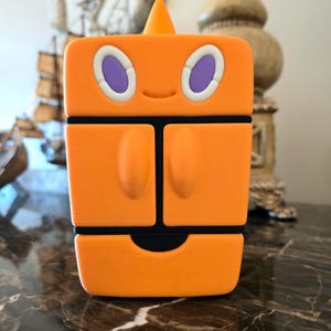 May include: A bright orange, rectangular, stylized character figure with a small pointed top, two oval purple eyes with white borders, and a subtle smile. The figure features a segmented design with two vertical panels and a horizontal base section.