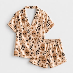 May include: A two-piece pyjama set featuring a short-sleeved button-down shirt and matching shorts. The fabric is a light beige colour with a repeating pattern of brown skulls, dark brown leafy branches, small hearts, and pale floral outlines.